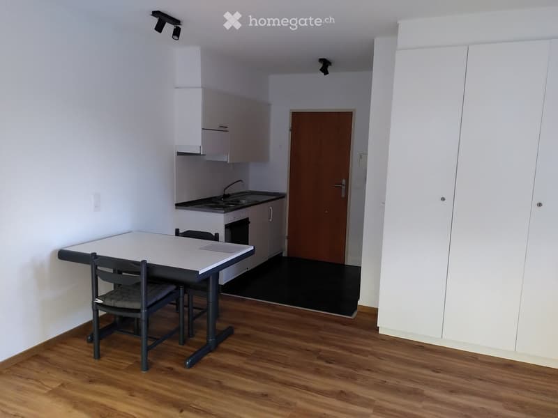 A compact, modern studio apartment with a wooden floor, white walls, a small kitchen area with cabinets and a sink, and a dining table with chairs.