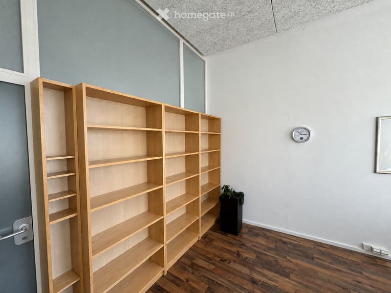 Empty room with wooden floor, light blue and white walls, large wooden shelving unit, small black speaker, wall clock, and a small potted plant near the shelving unit.