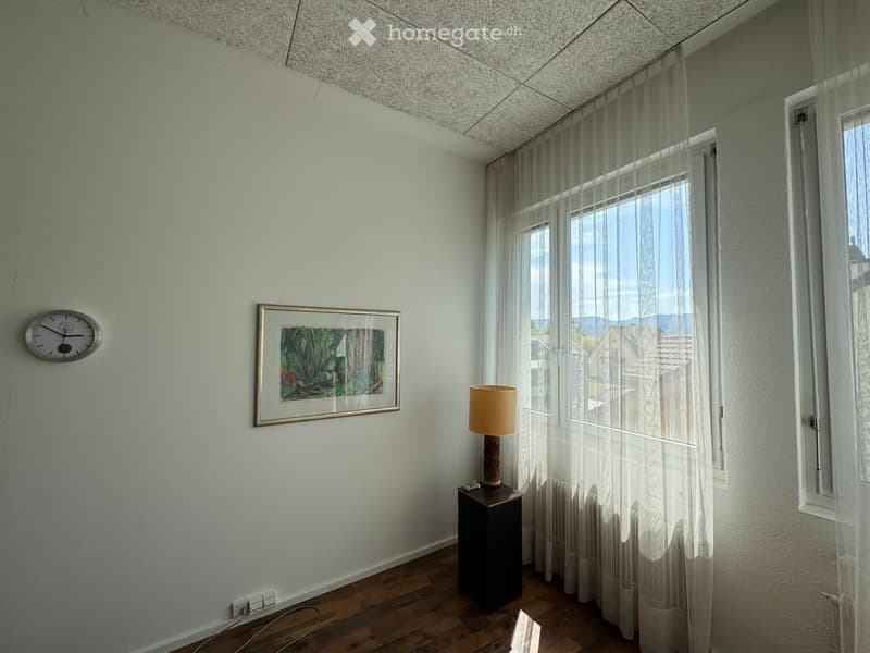 A small, well-lit room with wooden flooring, white walls, and large windows covered with sheer curtains. A small table with a lamp, a picture frame on the wall, and a wall-mounted clock are visible.