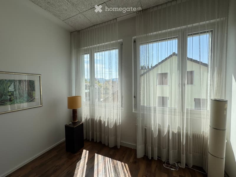 Empty room with wooden flooring, large windows with sheer curtains, a small side table with a lamp, and a framed picture on the wall. Natural light streams in, illuminating the space.
