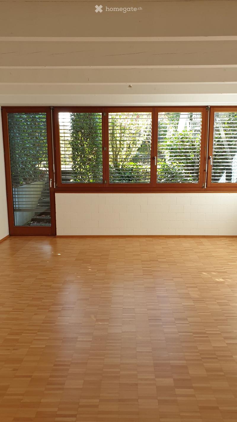 Empty room with wooden flooring and large windows with blinds, featuring white brick lower walls and a wooden door.