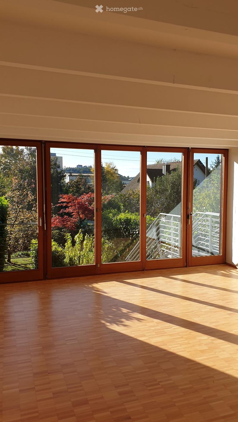 Empty room with wooden flooring and large sliding glass doors leading to a balcony or outdoor area. Sunlight streams in, casting shadows on the floor.