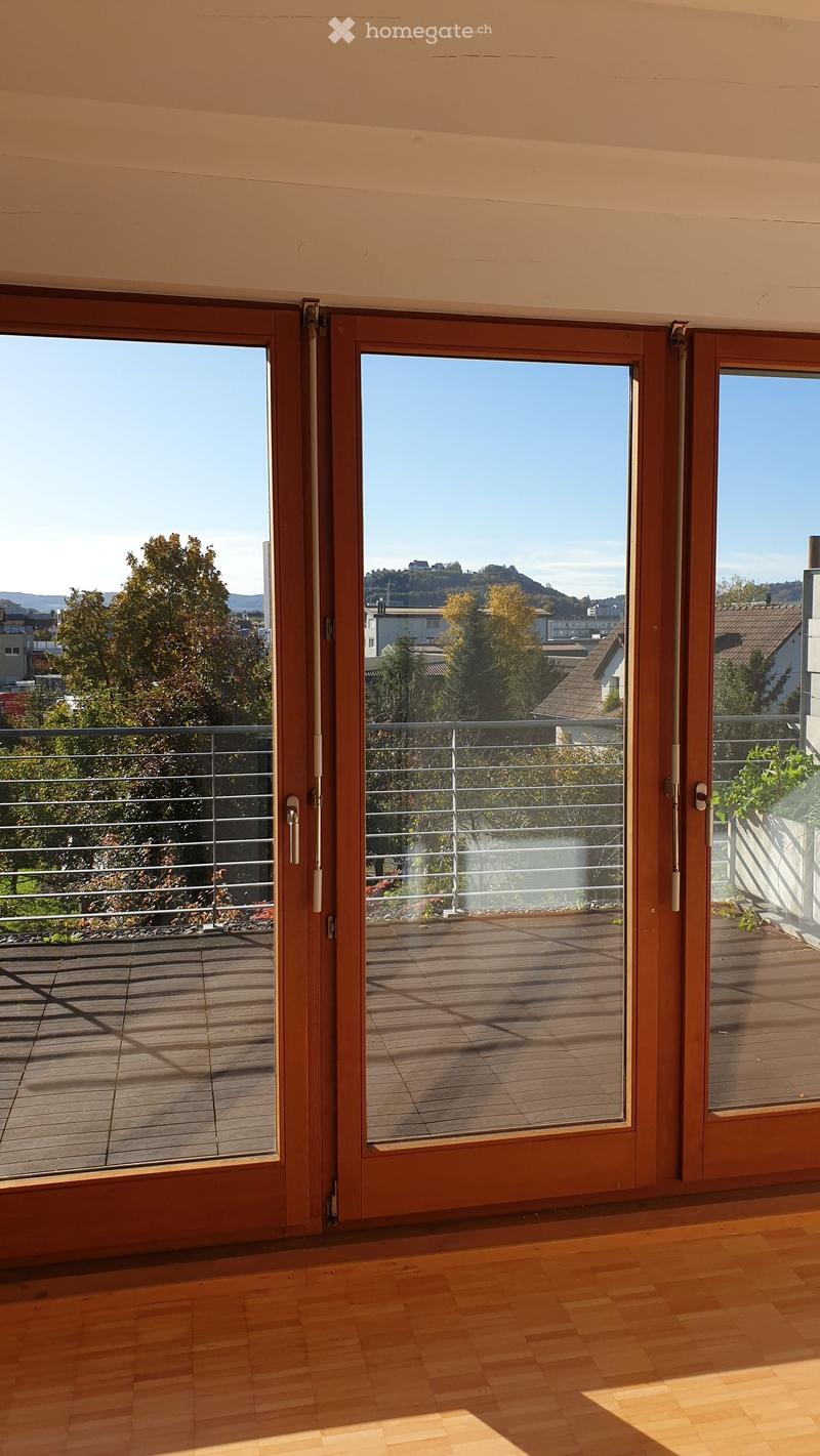Empty room with wooden flooring and large sliding glass doors leading to a wooden decked balcony with metal railings, overlooking a scenic view of trees and buildings.