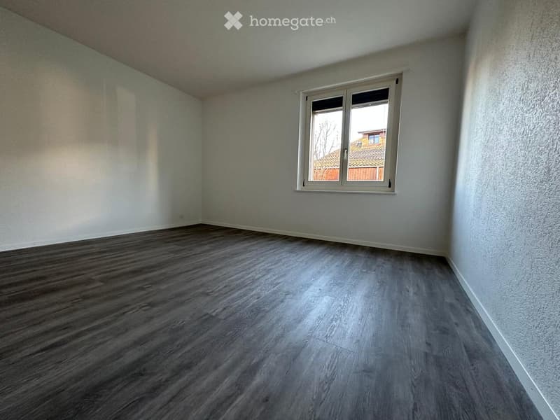 Empty room with light grey walls, wooden flooring, and a single window with white frame showing a view of a neighboring building and yard.