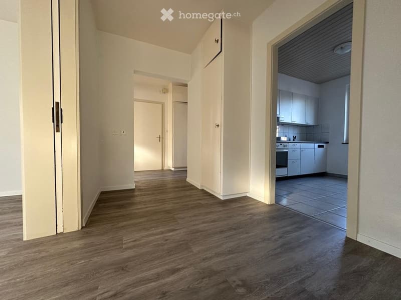 Empty hallway with wooden flooring, white walls, and doors leading to other rooms, including a kitchen with cabinets and appliances.