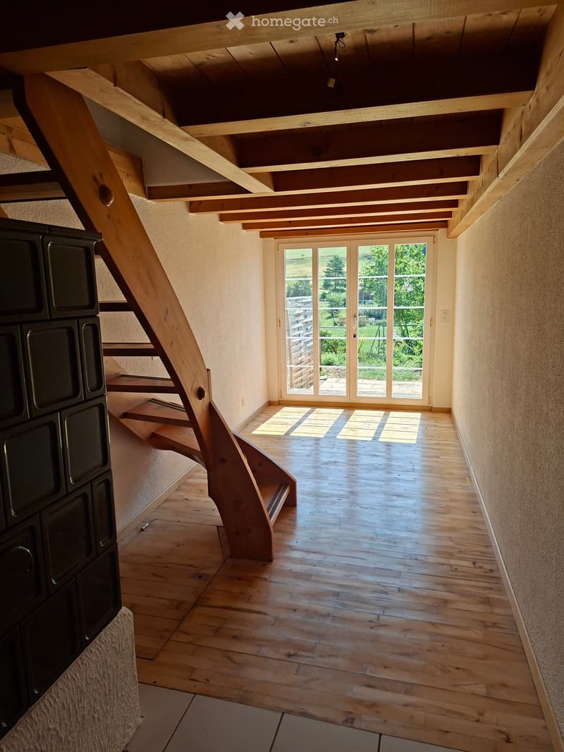 Bright, open hallway with wooden flooring, exposed wooden ceiling beams, and a curved staircase leading upwards. Large glass doors at the end provide natural light and a view of the outdoor area.