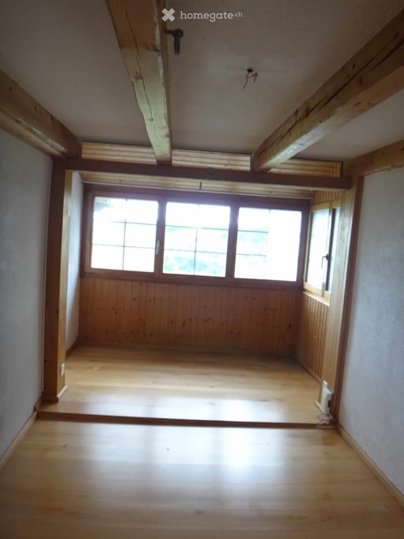 Empty room with wooden flooring, exposed wooden beams, and large windows allowing natural light.