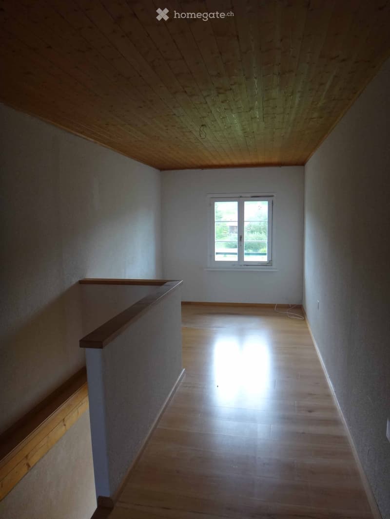 Empty hallway with wooden flooring, white walls, and a window at the end allowing natural light to enter.