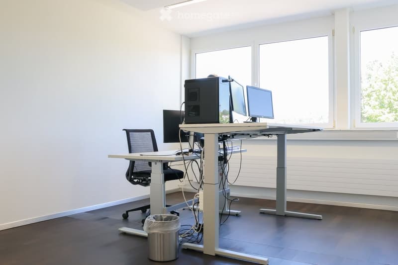 Modern office space with a standing desk, ergonomic chair, computer setup, and large windows providing natural light.