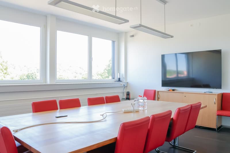 Bright conference room with large windows, a long wooden table surrounded by red chairs, a flat-screen screen mounted on the wall, and a small wooden cabinet.
