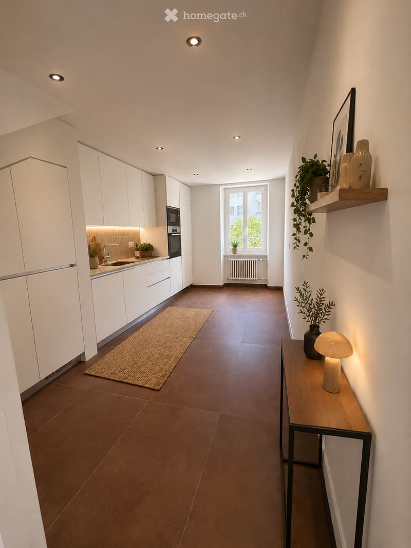 Modern hallway with brown tiled floor, white walls, built-in white kitchen cabinets on one side, and a small wooden table with a lamp and plant on the other side. A window at the end provides natural light.