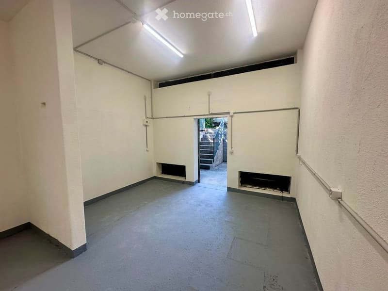Empty hallway with white walls, grey concrete floor, fluorescent lighting, and two black vents on the walls. An open doorway leads to a staircase outside.