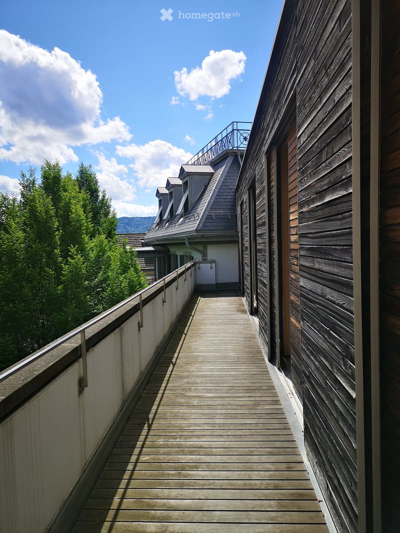 Wooden balcony with metal railing, overlooking a green landscape and a building with a grey roof.