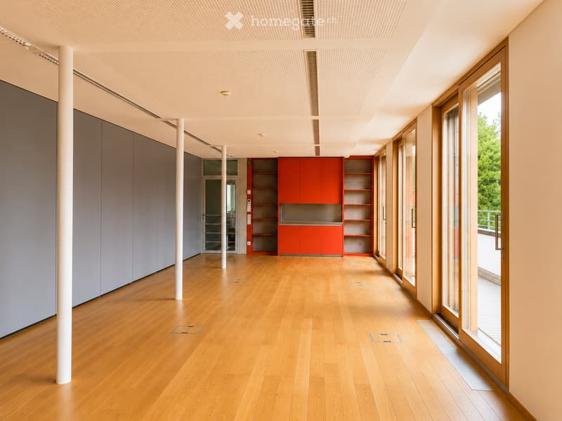 Empty room with wooden flooring, white walls, and large windows. Features include a red cabinet, white columns, and a sliding glass door leading to a balcony.