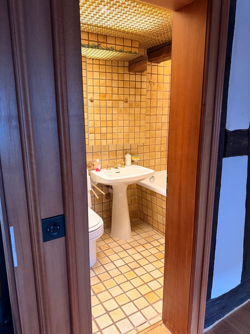 Small bathroom with beige tiles, a toilet, a pedestal sink, and a bathtub. Features a decorative ceiling with a gold-patterned design and a small shelf above the sink.