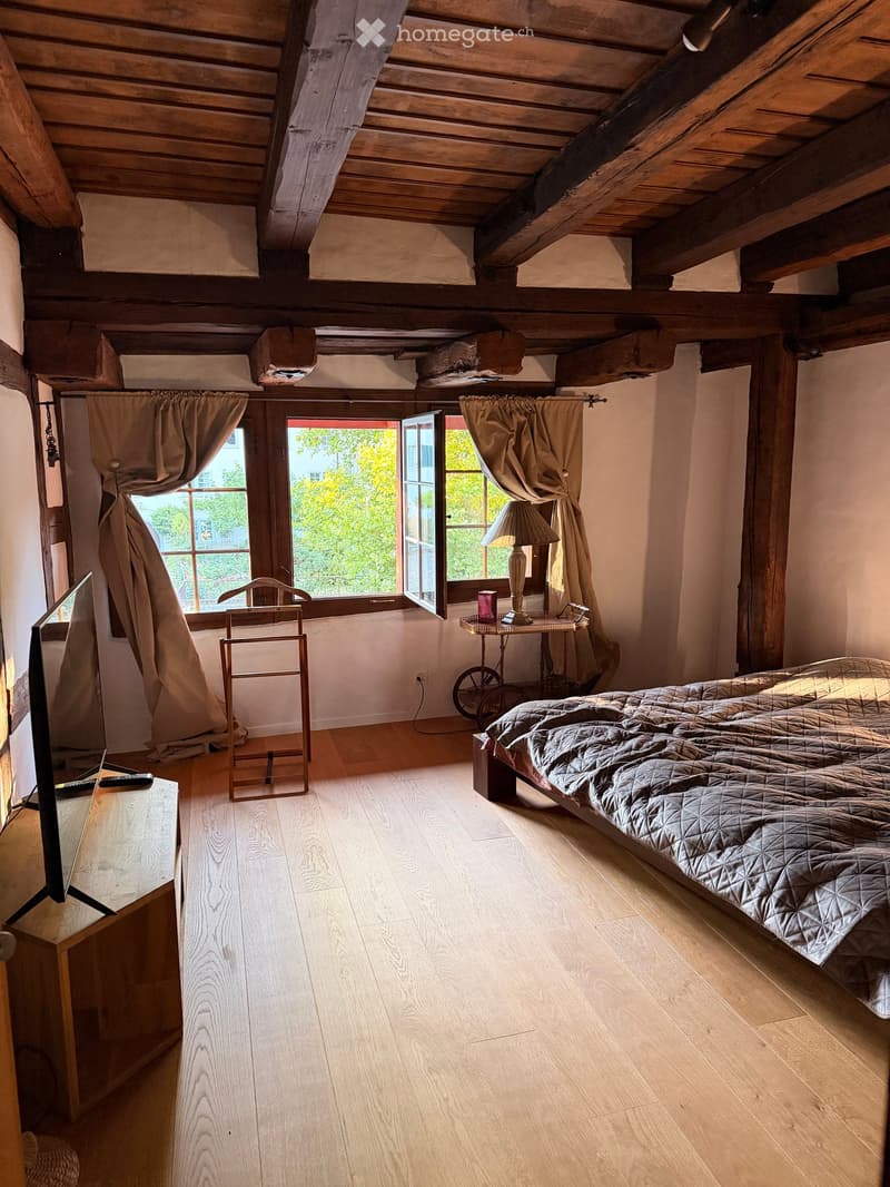 Cozy attic bedroom with wooden beams, large windows, a bed with a quilted cover, a small table with a lamp, and a chair near the window.