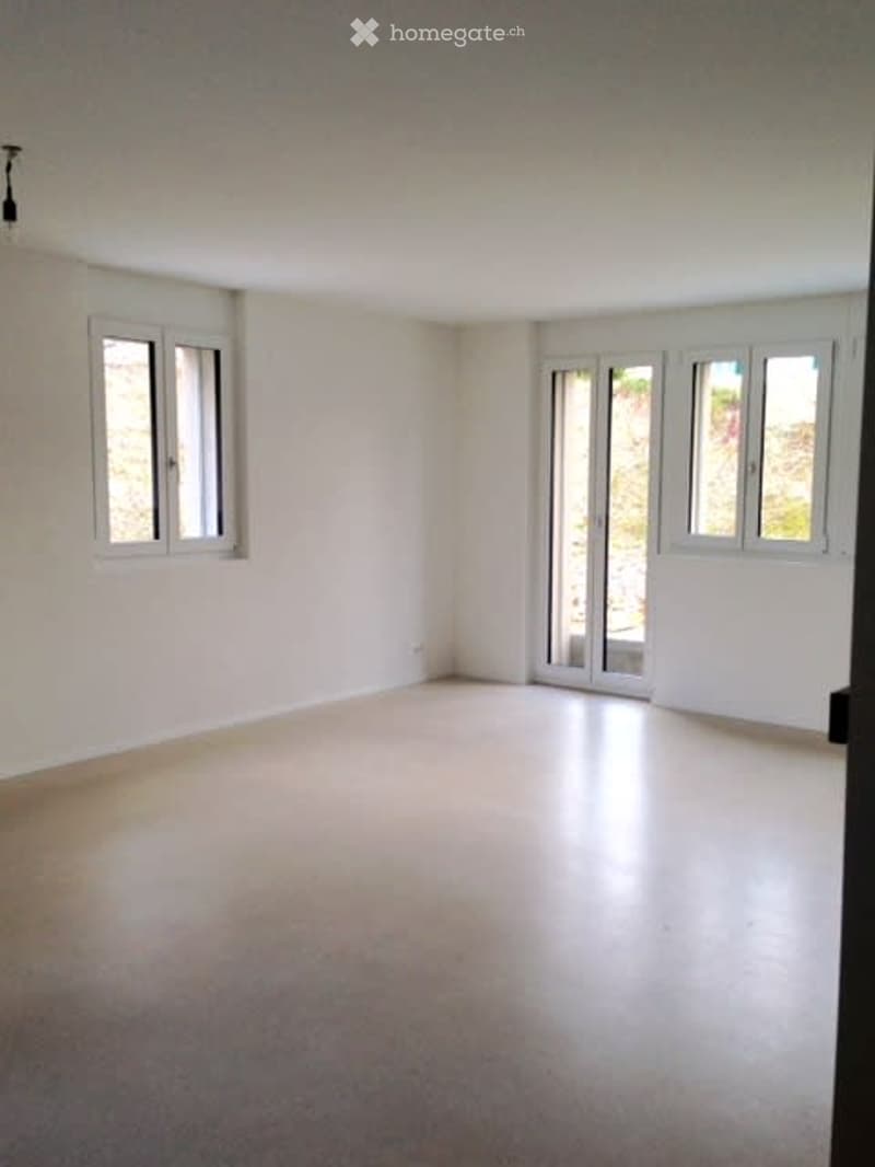 Empty room with white walls, large windows, and a sliding glass door leading to an outdoor area. The floor is a smooth, light-colored material.