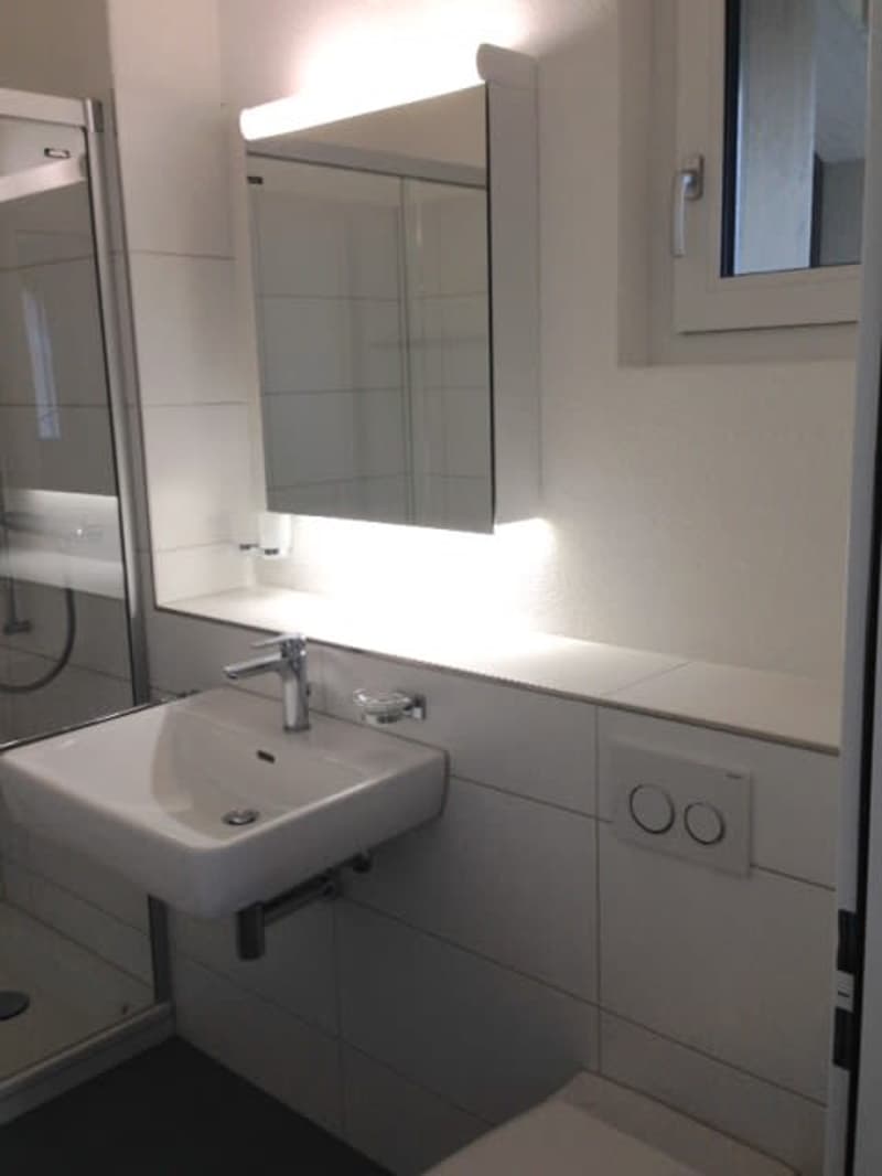 Modern bathroom with a white sink, chrome faucet, and a large mirror above it. The walls are tiled in light gray, and there is a small window on the right side.