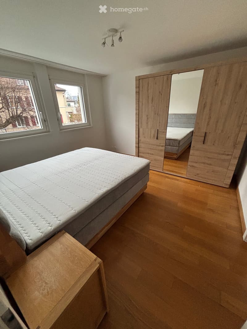Modern bedroom with wooden flooring, large windows, a single bed with mattress, a wooden wardrobe with mirrored doors, and a small wooden nightstand.