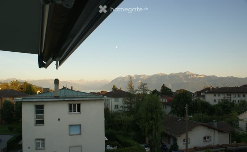 Balcony view overlooking a residential area with houses, trees, and distant mountains under a clear sky.