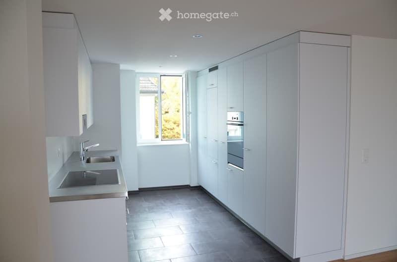 Modern, minimalist kitchen with white cabinets, stainless steel sink, and built-in oven. Features a large window with a view of greenery outside.