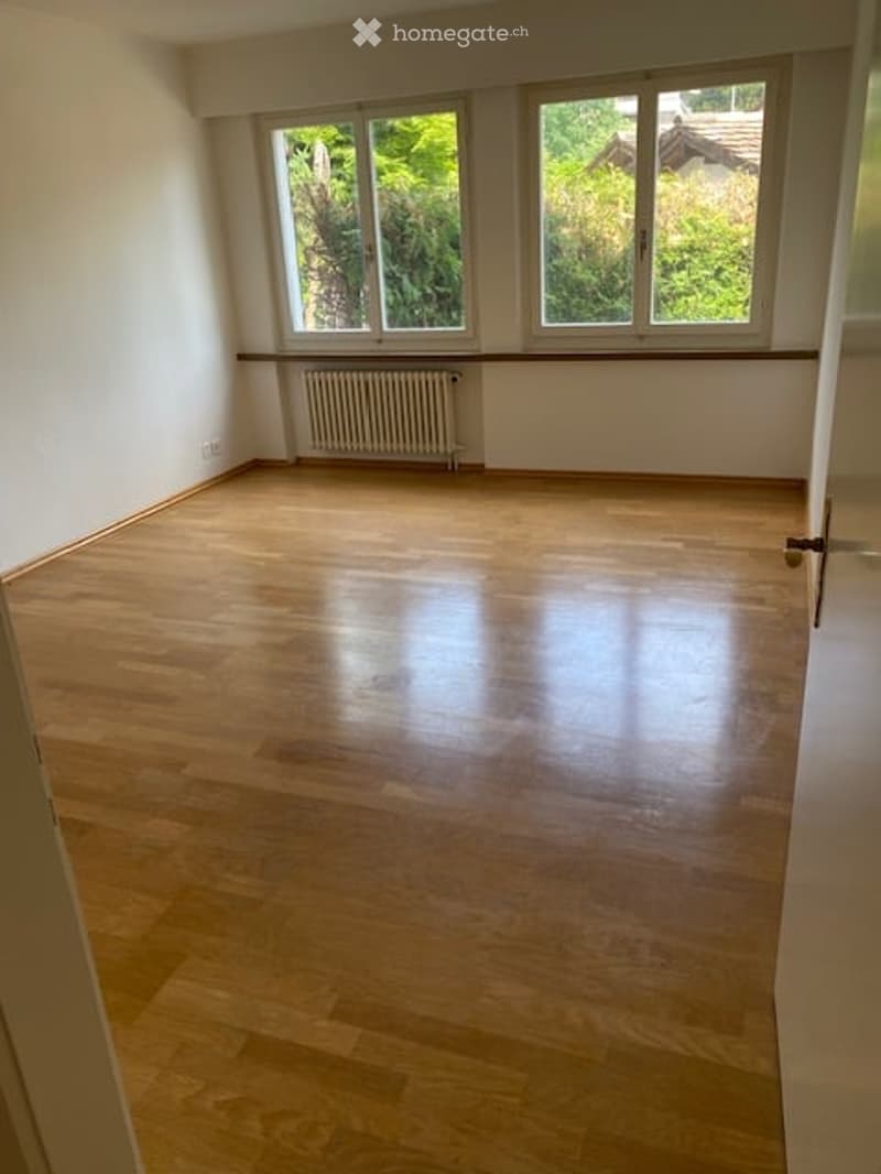 Empty room with wooden flooring, white walls, and large windows with white frames. A radiator is visible under the windows.