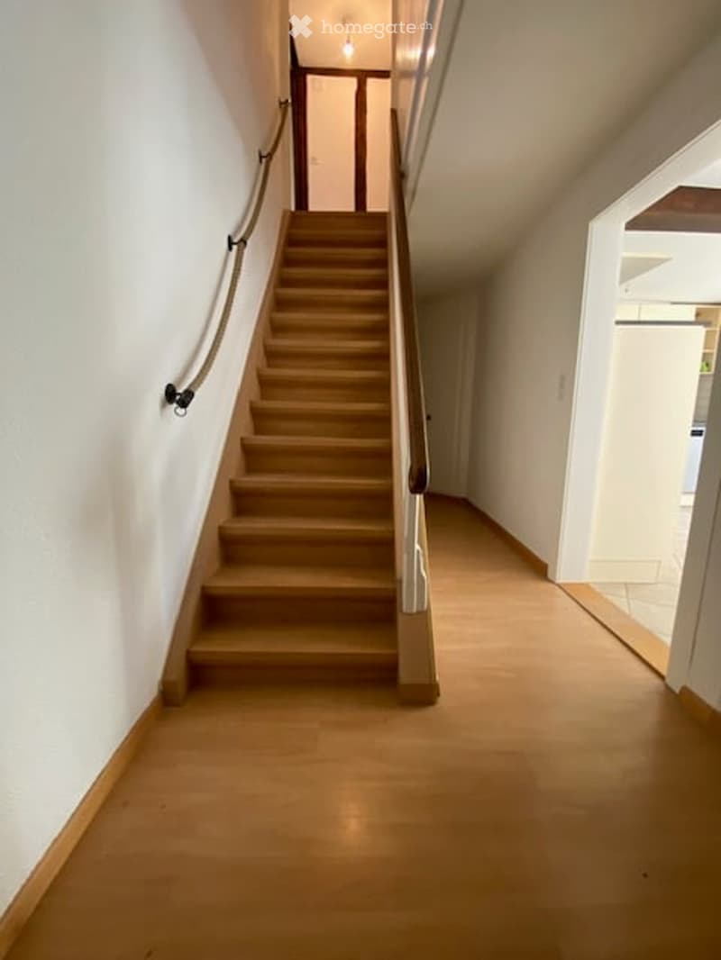 Spacious hallway with wooden flooring and a staircase leading upwards, featuring a handrail and a light fixture at the top.