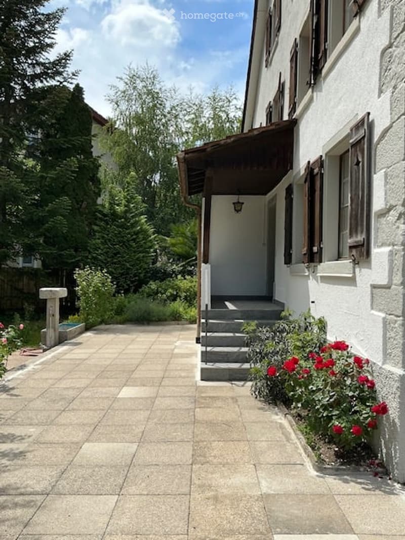 A white building with a covered entrance, steps leading up to the door, and a small garden area with red flowers and greenery.