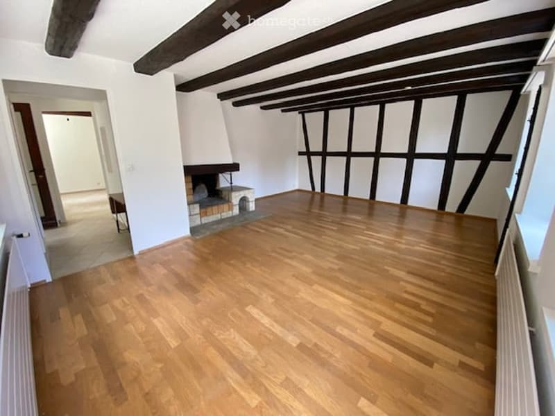 Empty room with wooden flooring, exposed wooden beams, white walls, and a fireplace. No furniture or equipment present.