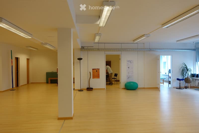 Open-plan room with light wooden flooring, white walls, and modern lighting fixtures. Features include a large green ball, a small wooden stool, a tall floor lamp, and several chairs and tables in the background.