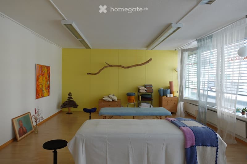 Brightly lit room with wooden flooring, white walls, and a yellow accent wall. Features a massage table, a wooden bench, a small shelf with folded towels, a stool, and decorative items including a framed picture and a vase with dried flowers.