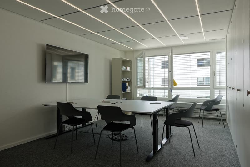 Modern meeting room with a large rectangular table, six chairs, a wall-mounted screen, and a small shelf with books and office supplies. Large windows with blinds allow natural light to fill the space.