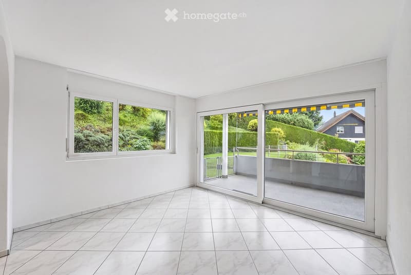 Bright, empty room with white tiled flooring and white walls. Features large windows and sliding glass doors leading to a balcony or terrace area with a view of greenery and a neighboring house.