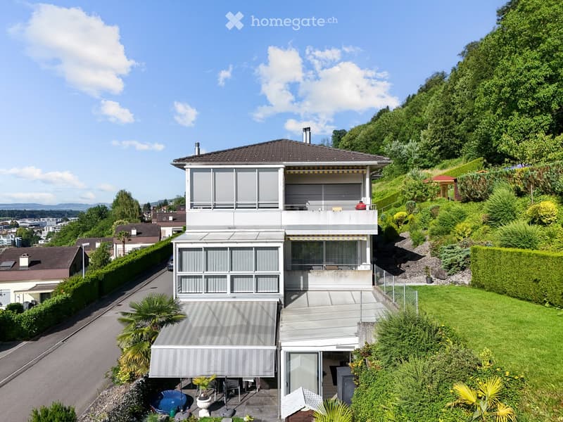 Modern two-story house with large windows, balconies, and a well-maintained garden with palm trees and a small swimming pool.