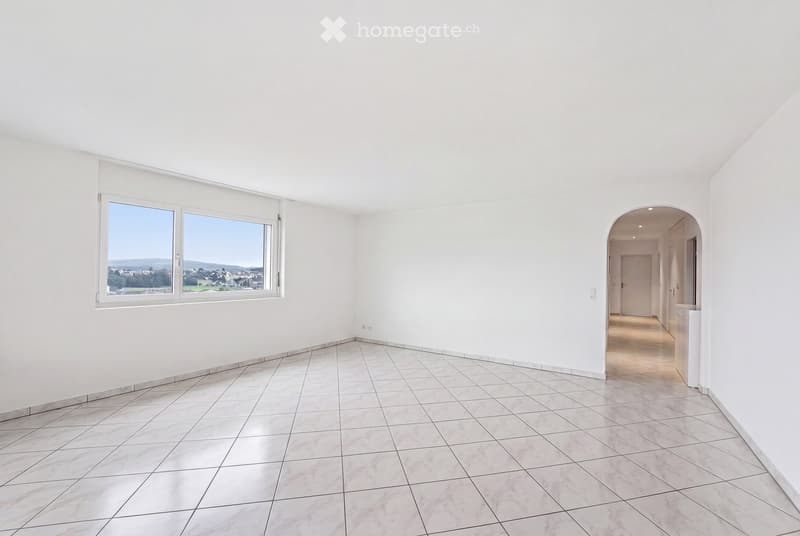 Empty room with white walls, tiled floor, large window with blinds, and an arched doorway leading to another area.