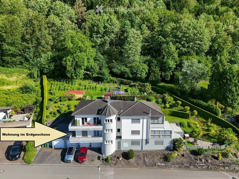 A modern two-story house with a raised ground floor, surrounded by lush greenery and trees. The house features a garage, multiple balconies, and a well-maintained garden area.
