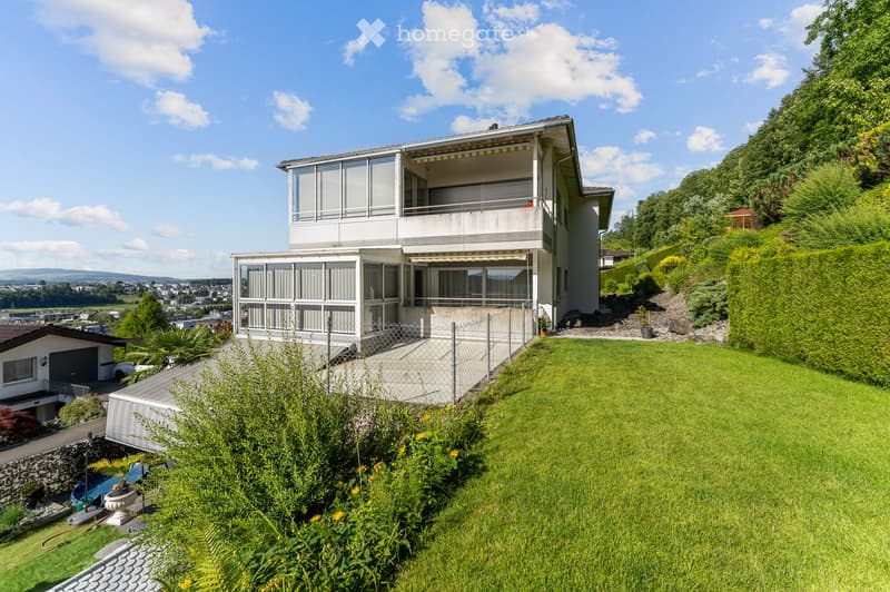 A modern two-story house with large windows and balconies, situated on a hillside with a well-maintained lawn and surrounding greenery.