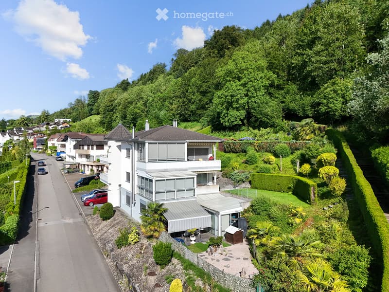 A two-story house with a modern design, featuring a balcony, garage, and well-maintained garden with various plants and trees.
