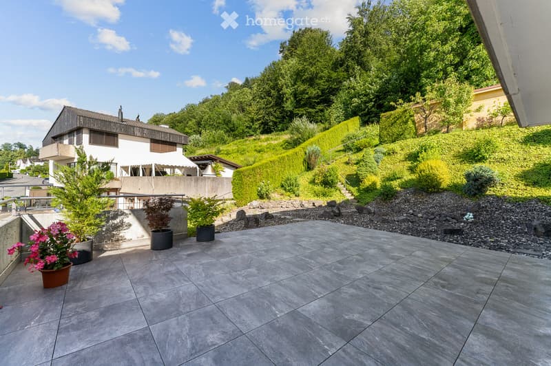 A spacious outdoor patio with a tiled floor, surrounded by greenery and potted plants, with a modern house and hillside in the background.