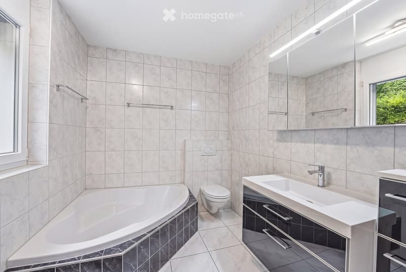Modern bathroom with a large bathtub, toilet, double sink, and large mirror. The walls are tiled in a light marble pattern, and the floor is tiled in a dark marble pattern. There are windows on two sides, allowing natural light to enter.