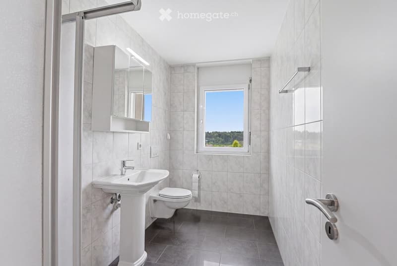 Modern bathroom with white tiles, a sink, toilet, mirror, and a window offering a view of greenery.