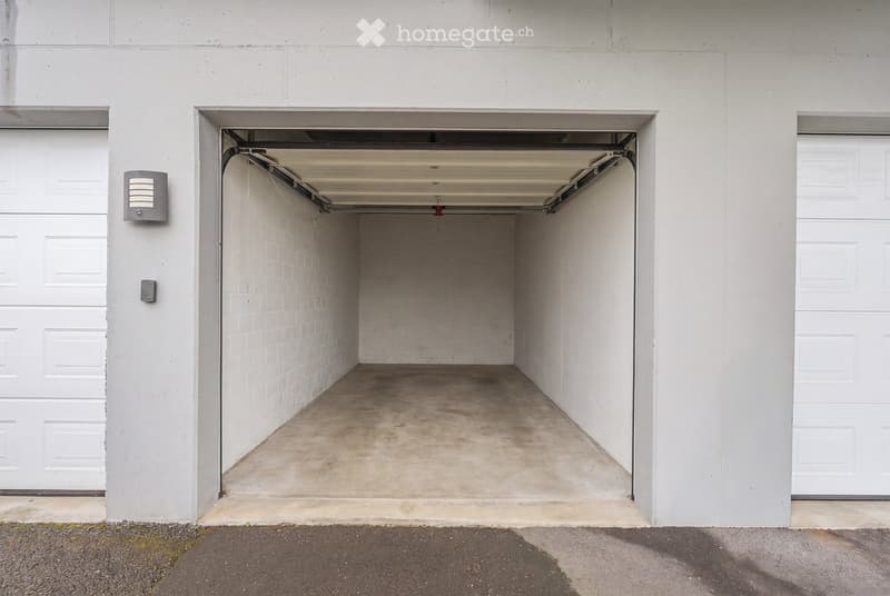 A clean, empty garage with white walls, a concrete floor, and overhead lighting. The garage door is open, revealing the space.
