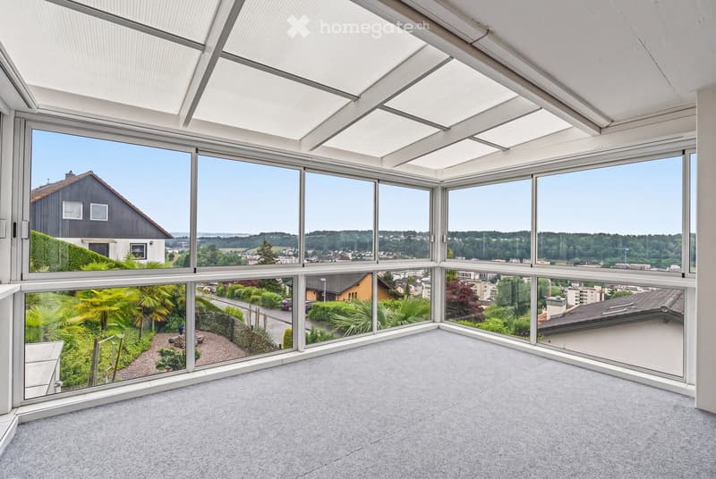 Bright, spacious room with large windows offering panoramic views of a town and surrounding landscape. The room features a high, slanted ceiling and a carpeted floor.