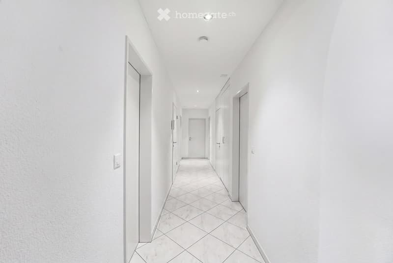 A clean, modern hallway with white walls, tiled floor, and several closed doors along both sides. Recessed ceiling lights provide illumination.