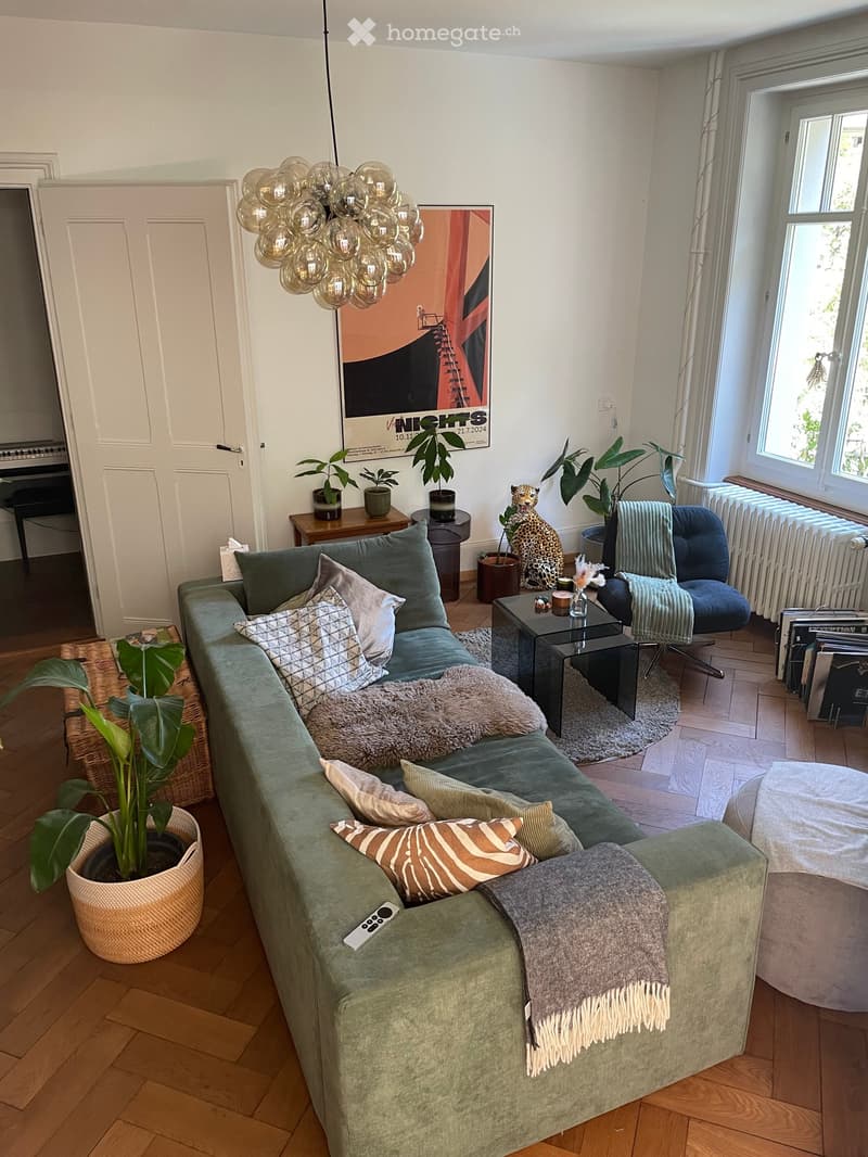 A modern living room with a green sectional sofa, multiple throw pillows, a coffee table, a small side table, a large floor lamp, and several potted plants. The room features wooden flooring and large windows letting in natural light.