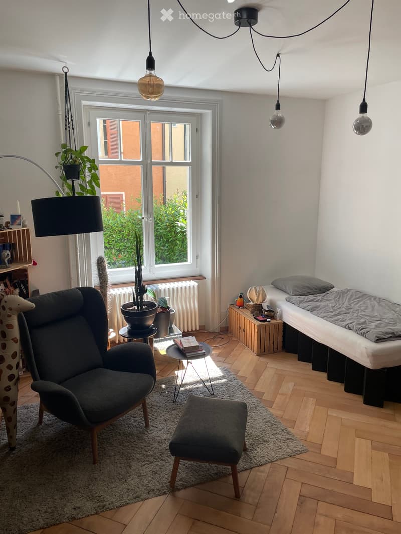Cozy bedroom with wooden flooring, large window with view, sofa, stool, small table, bed, and decorative items.