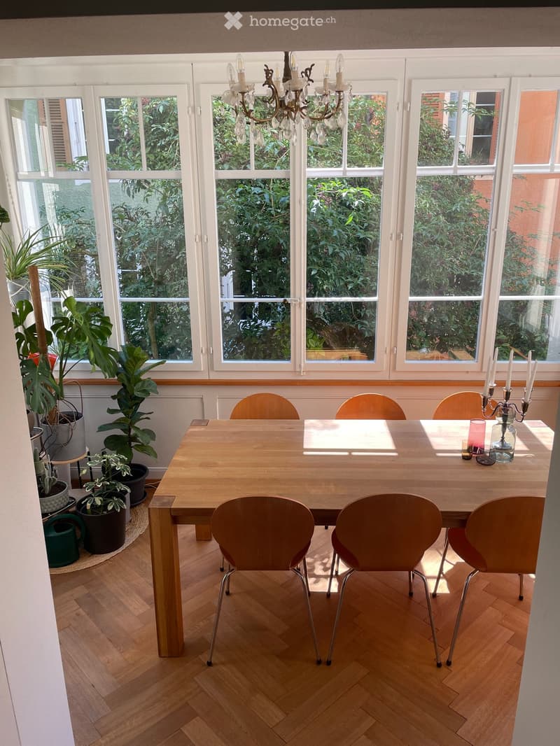 Brightly lit dining room with large windows, wooden dining table, six chairs, candle holder with candles, and several potted plants.