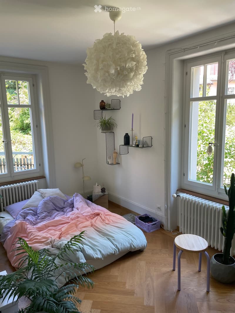 A cozy bedroom with a large bed covered in pastel-colored bedding, a wooden floor, and large windows letting in natural light. The room features a hanging decorative light fixture, a small table with a lamp, shelves with decorative items, and potted plant