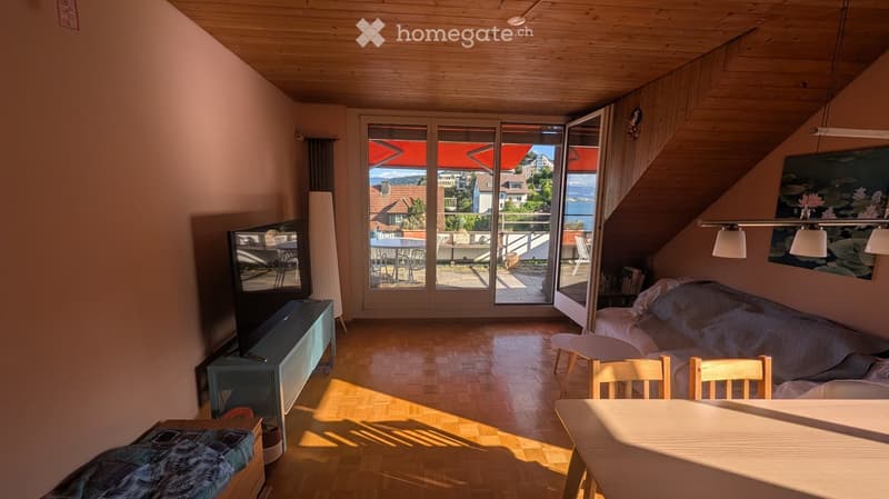 Bright living room with wooden floors, large windows, and a view of the lake and surrounding houses. Features a sofa, dining table, chairs, and a television.
