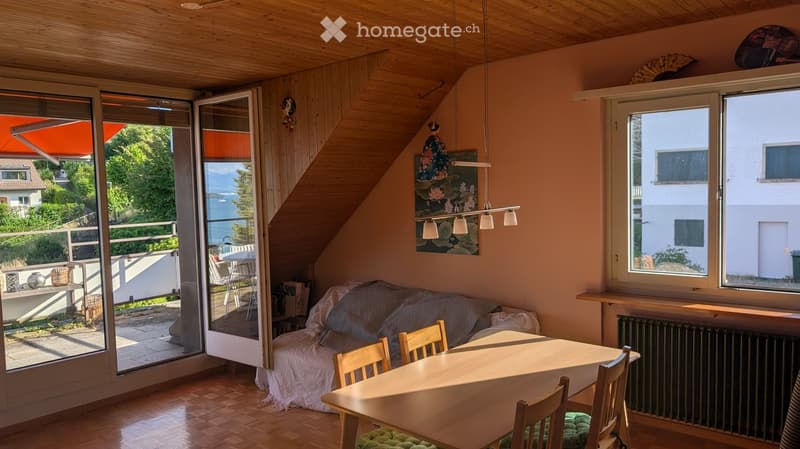 Bright living room with wooden floors, large windows, and a view of the lake. Features a sofa, dining table with chairs, radiator, and artwork on the wall.